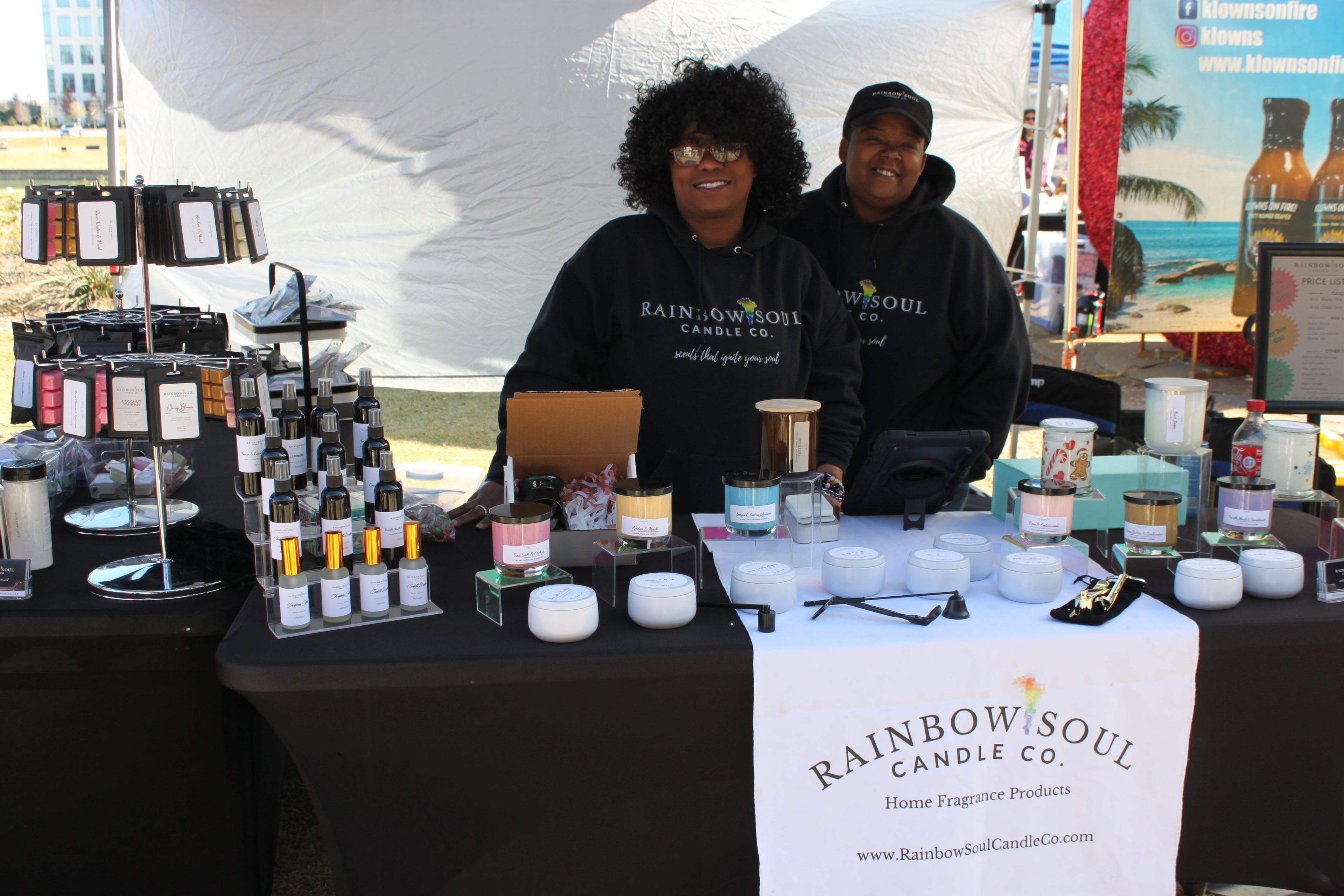 Two individuals behind a table displaying products with a 'Rainbow Soul Candle Co.' banner.